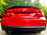 A red BMW M235i features prominently, parked on a gravel surface, surrounded by lush greenery. The BMW and M235i logos are visible on the back, with dual exhausts below.