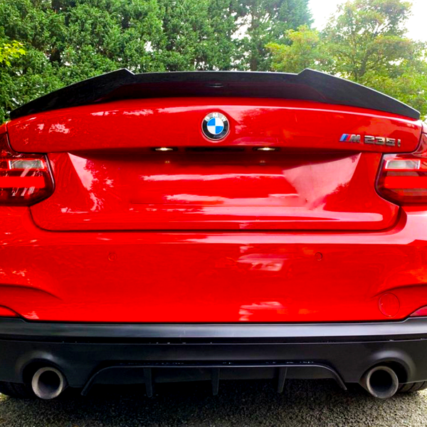 A red BMW M235i features prominently, parked on a gravel surface, surrounded by lush greenery. The BMW and M235i logos are visible on the back, with dual exhausts below.