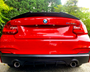 A red BMW M235i features prominently, parked on a gravel surface, surrounded by lush greenery. The BMW and M235i logos are visible on the back, with dual exhausts below.