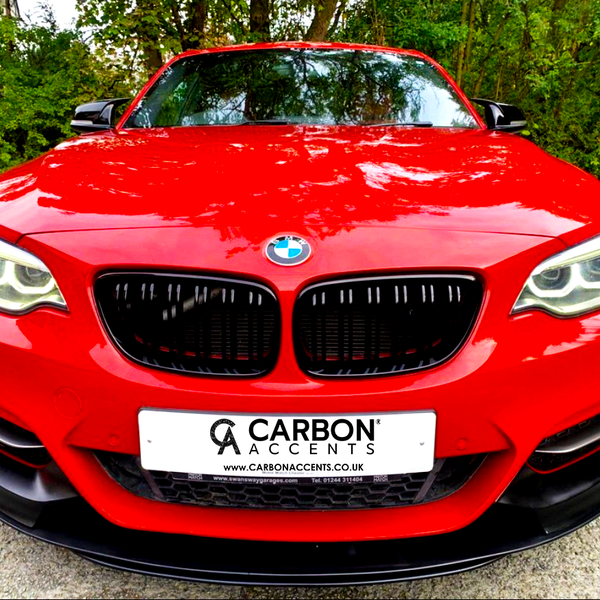 A red BMW car is parked on a concrete surface, front view, with trees in the background. The license plate reads, "CARBON ACCENTS www.CARBONACCENTS.co.uk."