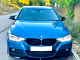 A blue BMW is parked on a roadside, headlights on. Surrounding it are trees and bushes, with a wooden fence along the right side.