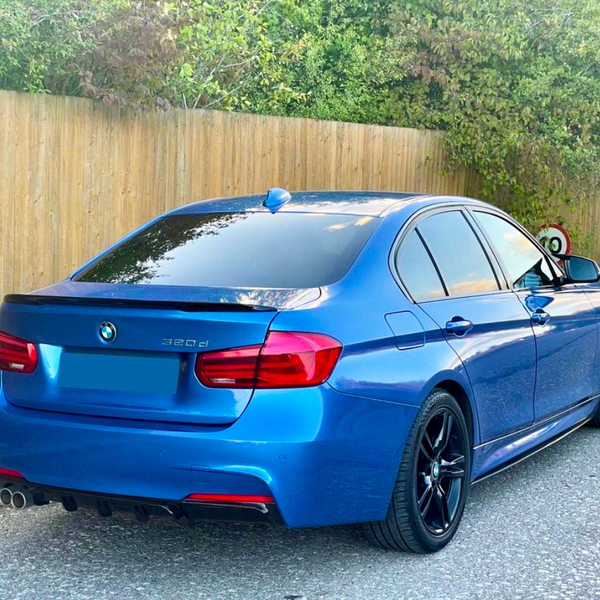A blue BMW 320d is parked on a paved street next to a wooden fence, surrounded by greenery and a 20 mph speed limit sign.