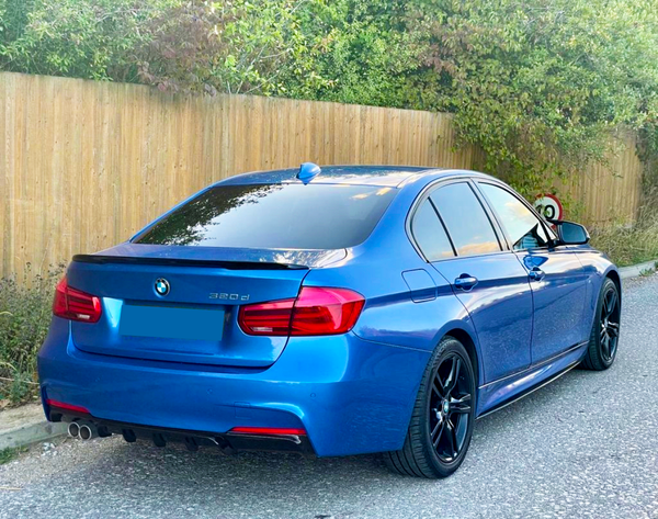 A blue BMW 320d is parked on a paved street next to a wooden fence, surrounded by greenery and a 20 mph speed limit sign.