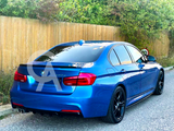 A blue BMW sedan is parked on a paved road, facing away, near a wooden fence with greenery. The model number 