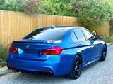 A blue BMW 320d is parked on a road, facing away. It features tinted windows and black wheels, adjacent to a wooden fence and greenery.