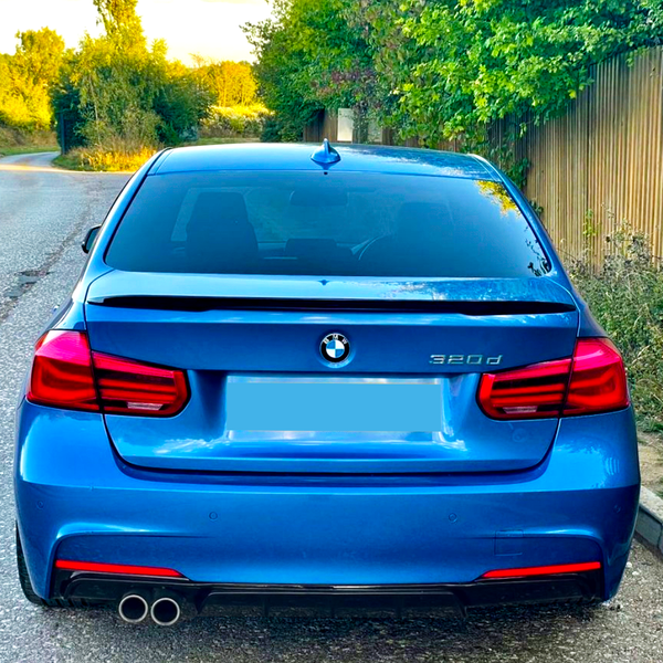 A blue BMW 320d is parked on a country road. Trees and a wooden fence line the roadside, creating a serene, rural environment.