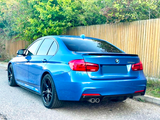 A blue BMW 320d parked on a road beside a wooden fence and trees, showcasing its rear with dual exhausts and tinted windows.
