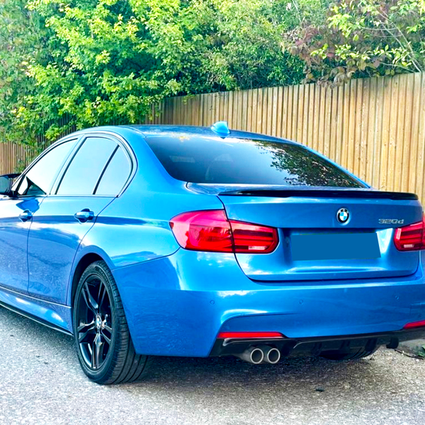 A blue BMW 320d parked on a road beside a wooden fence and trees, showcasing its rear with dual exhausts and tinted windows.