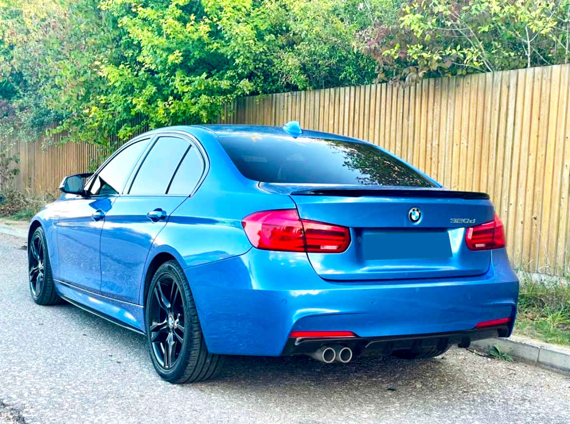 A blue BMW 320d parked on a road beside a wooden fence and trees, showcasing its rear with dual exhausts and tinted windows.