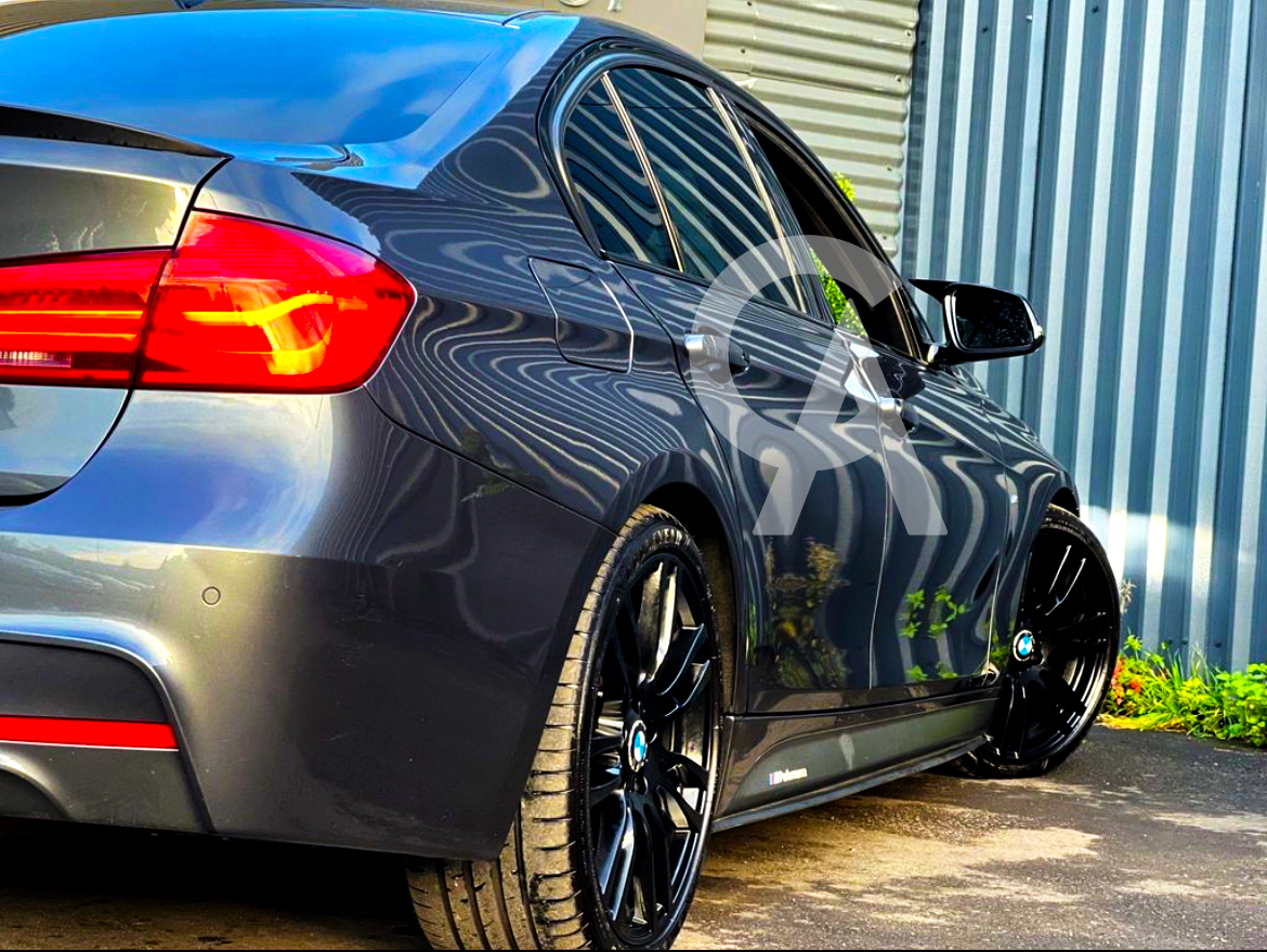 A sleek, grey sedan with tinted windows and bright red taillights is parked on pavement, reflecting striped patterns from a nearby corrugated metal wall and surrounded by green foliage.