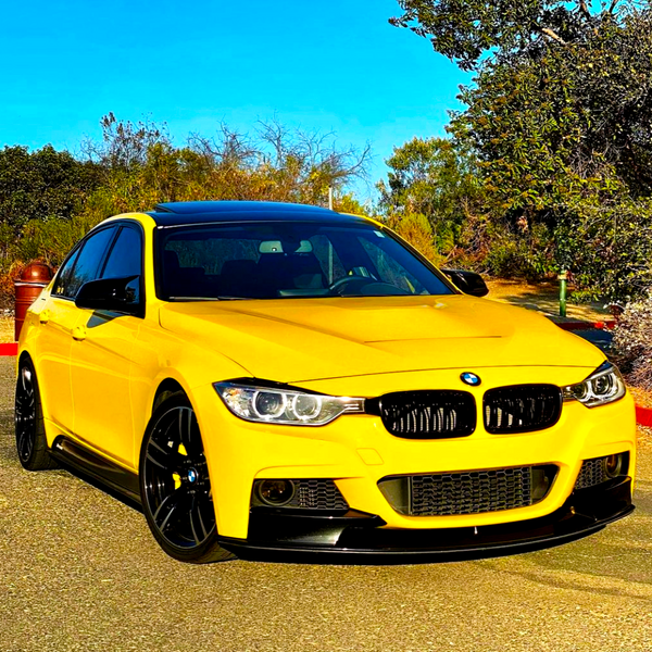 A yellow BMW car is parked, facing forward on a sunny driveway, surrounded by trees and shrubs under a clear blue sky.