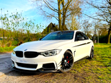 A white sports car with sleek black rims is parked on dirt, surrounded by trees and greenery under a clear blue sky.