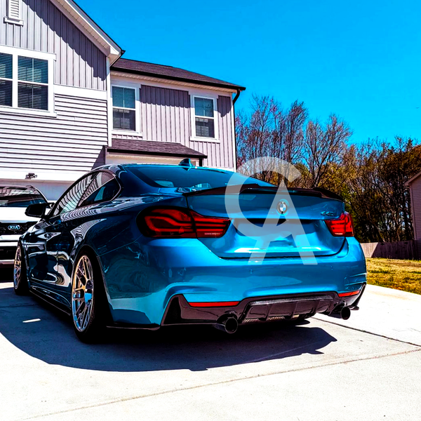 Blue BMW parked on a driveway, facing away from a two-story house. A white Kia is parked nearby, with trees and a blue sky in the background.