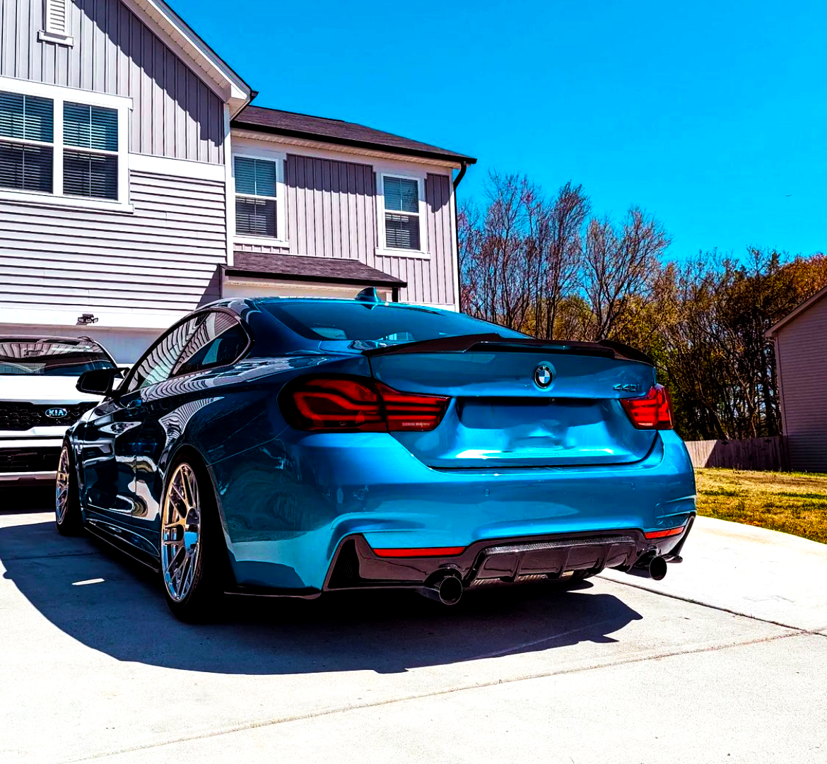 A bright blue car is parked in a driveway, facing a two-story house. Another vehicle, partially visible, is parked beside it. Trees and a clear blue sky surround the scene.