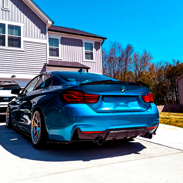 A bright blue car is parked in a driveway, facing a two-story house. Another vehicle, partially visible, is parked beside it. Trees and a clear blue sky surround the scene.