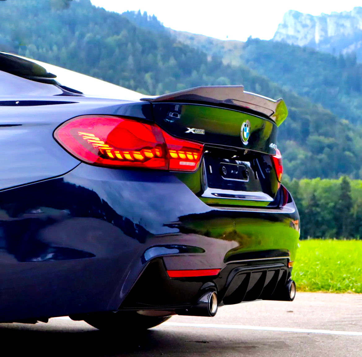 A dark blue BMW with illuminated taillights is parked on a paved road, surrounded by green fields and mountains. The car features dual exhausts. Visible text: "XDRIVE" and "430i."