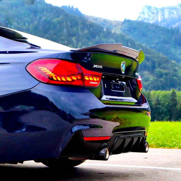 A dark blue BMW with illuminated taillights is parked on a paved road, surrounded by green fields and mountains. The car features dual exhausts. Visible text: "XDRIVE" and "430i."