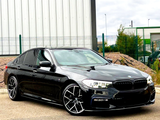 A sleek, black BMW car, parked with its headlights on, sits in an industrial area with a metal building and fences in the background.