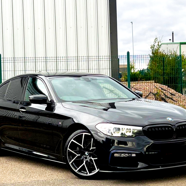 A sleek, black BMW car, parked with its headlights on, sits in an industrial area with a metal building and fences in the background.