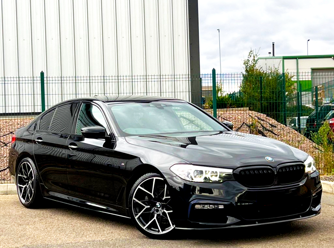 A sleek, black BMW car, parked with its headlights on, sits in an industrial area with a metal building and fences in the background.