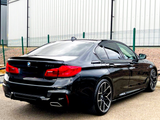 Black BMW sedan parked next to a fence, with shiny rims and distinctive red taillights, positioned on a concrete surface beside a large industrial-looking building.