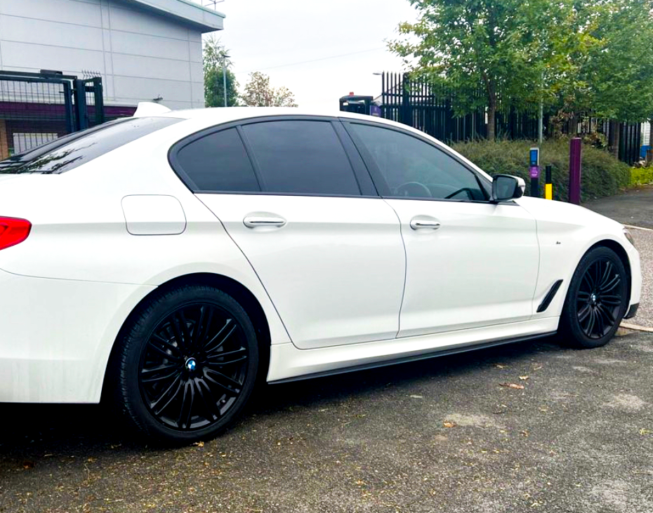 A white sedan with tinted windows and black wheels is parked on a paved surface. It is near a fenced area with trees and an industrial building in the background.