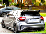 A silver sports hatchback with AMG and Mercedes badges, parked beside a road, features custom rims and a rear spoiler in a suburban neighborhood with trees and flowered gardens.