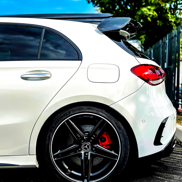 A white car is parked, showcasing its rear side with sleek black alloy wheels and a roof spoiler, amid a sunny day with a blue sky and some greenery nearby.