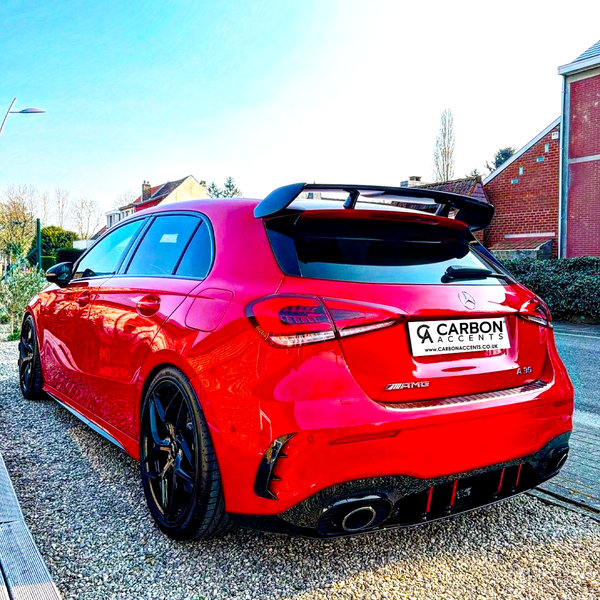 A red Mercedes-Benz parked on gravel, displaying a rear spoiler and dual exhausts, beside brick houses. License plate reads: "CARBON ACCENTS, WWW.CARBONACCENTS.CO.UK."