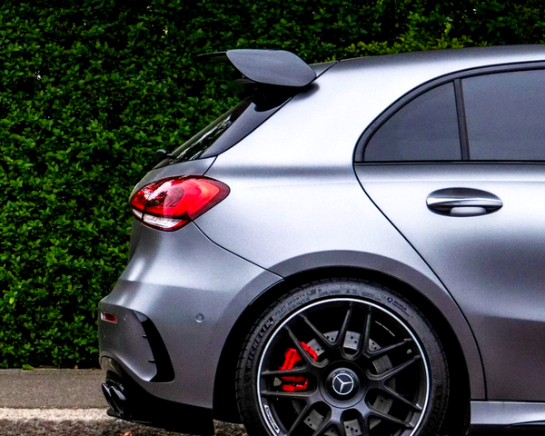 A silver car with a rear spoiler is parked beside a leafy hedge. Its red brake calipers are visible behind black alloy wheels.