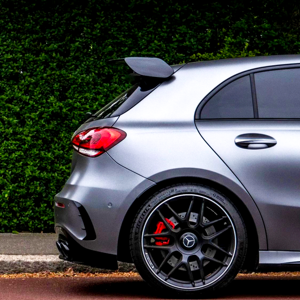 A silver car with a rear spoiler is parked beside a leafy hedge. Its red brake calipers are visible behind black alloy wheels.