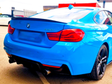 A blue BMW 440i car is parked on a light-colored surface, featuring dual exhausts and red-taillights. A fenced area and another parked car are visible in the background.