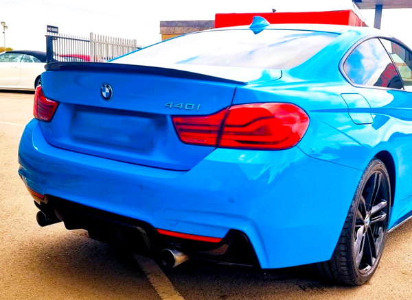 A blue BMW 440i car is parked on a light-colored surface, featuring dual exhausts and red-taillights. A fenced area and another parked car are visible in the background.