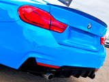 A blue BMW 440i with dual exhaust is parked on asphalt. Its distinctive rear lights and emblem are visible in bright daylight. Another car is partially visible nearby.