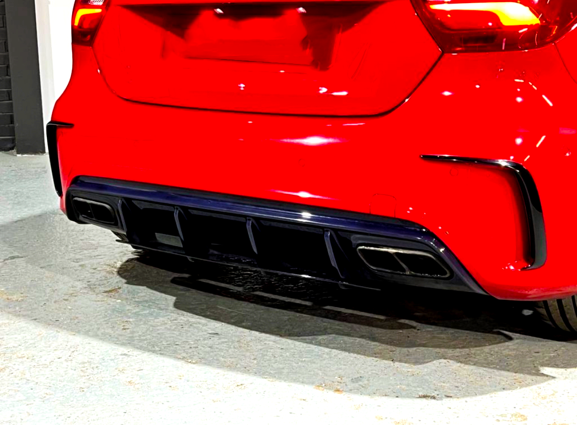 A red car's rear bumper features dual chrome exhausts and a black diffuser, parked on a light-colored concrete floor indoors.