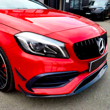 A red car with a shining grille and sleek headlights is parked outside a building on a paved surface. A black vehicle is visible in the background.