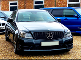 A black Mercedes-Benz is parked on gravel. It is flanked by a silver car on the left and a blue car on the right. The background features a brick building with windows.