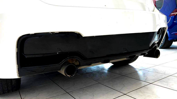 A black diffuser with dual exhaust pipes is attached to a white car, parked indoors on a tiled floor next to a blue vehicle.