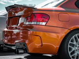 An orange BMW car is parked, showcasing its rear left side with tail lights, spoiler, and dual exhaust against a cloudy sky backdrop.