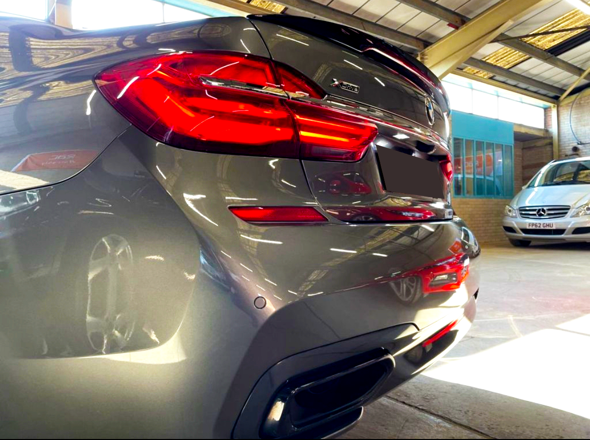 A gray BMW X5 car, parked indoors with illuminated brake lights, reflects its surroundings. Nearby, a silver Mercedes-Benz displays the license plate "FP62 GHU" in the bright-lit garage.