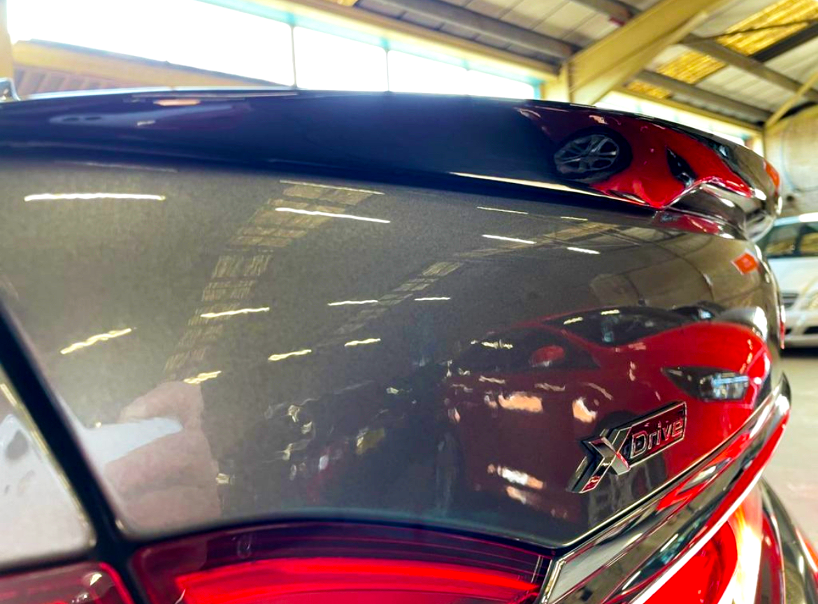 Close-up of a car's sleek, reflective surface displaying the "XDrive" badge, surrounded by reflections of red cars in a spacious, industrial garage with a high ceiling.