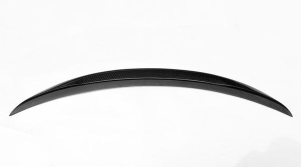 A sleek, curved car spoiler rests in a plain white environment, showcasing its black carbon-fiber texture prominently across its surface.