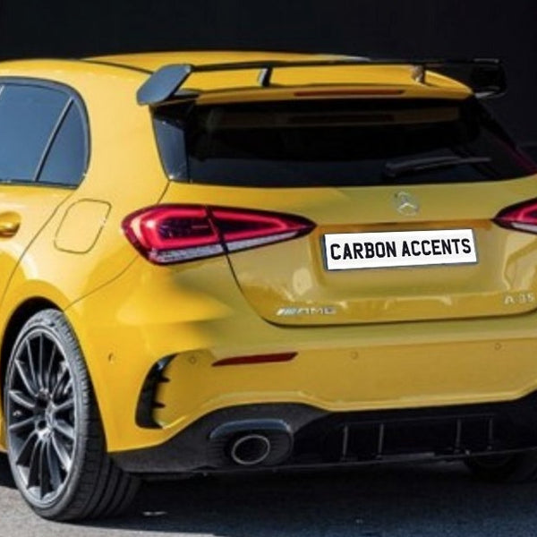 Yellow hatchback car parked, featuring a rear spoiler and dual exhaust. The license plate reads "CARBON ACCENTS." Vehicle is positioned near a red and black wall, suggesting an urban setting.
