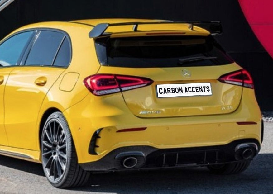 Yellow hatchback car parked, featuring a rear spoiler and dual exhaust. The license plate reads "CARBON ACCENTS." Vehicle is positioned near a red and black wall, suggesting an urban setting.