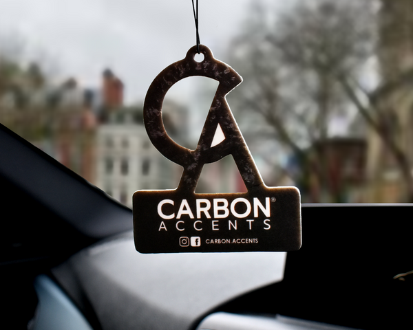 Air freshener hangs in a car, featuring the logo "CA" with text "CARBON ACCENTS" and "IG FB: @CARBON.ACCENTS." Blurred city buildings and trees visible through the windshield.