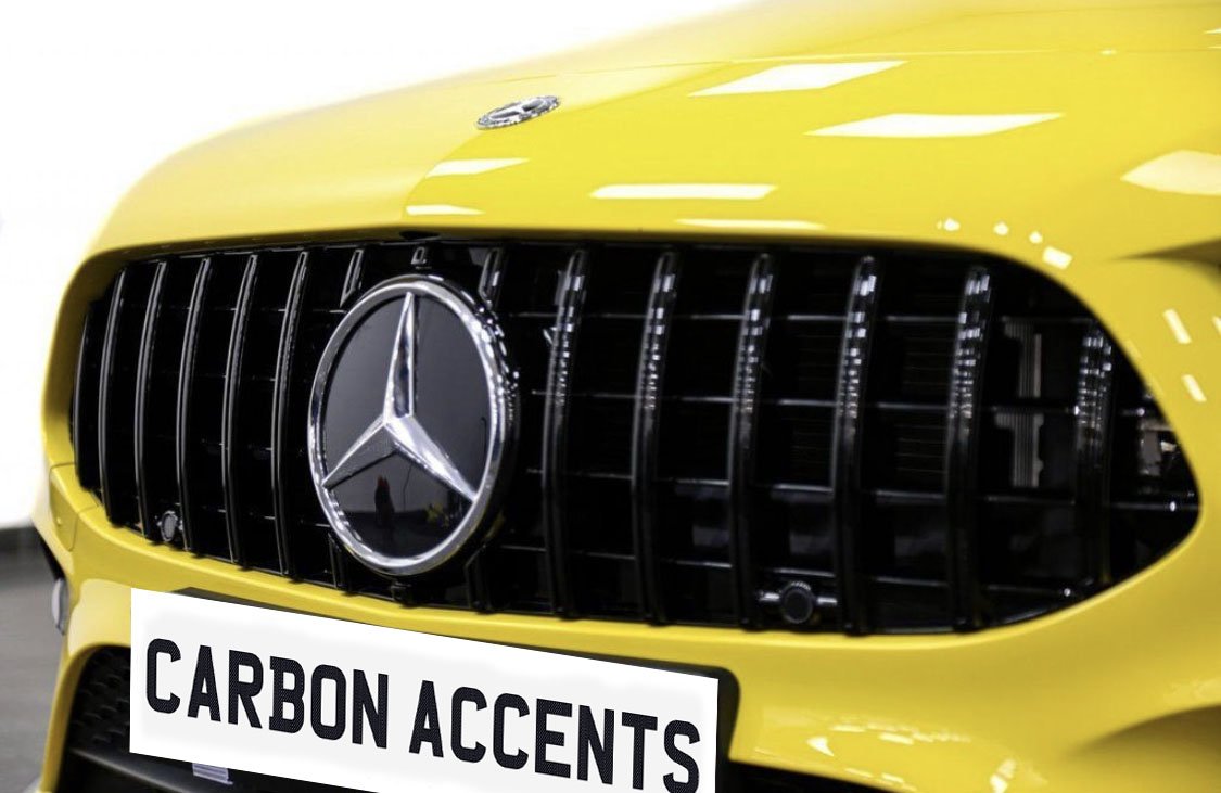 A yellow car's front grille features a prominent emblem and vertical bars. Below, a license plate reads "CARBON ACCENTS." The background is an indoor setting.