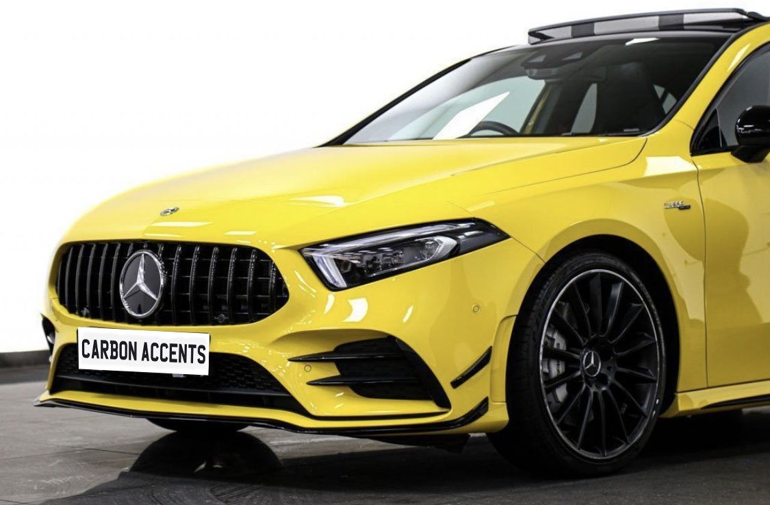 Yellow luxury car with a prominent logo is parked on a showroom floor. The license plate reads "CARBON ACCENTS." The car features sleek black wheels and a modern design.