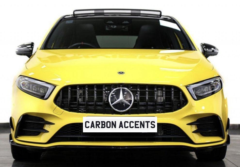 A bright yellow car with a prominent front grille displays a logo. Text on the front reads "CARBON ACCENTS." It's situated indoors on a plain white background.