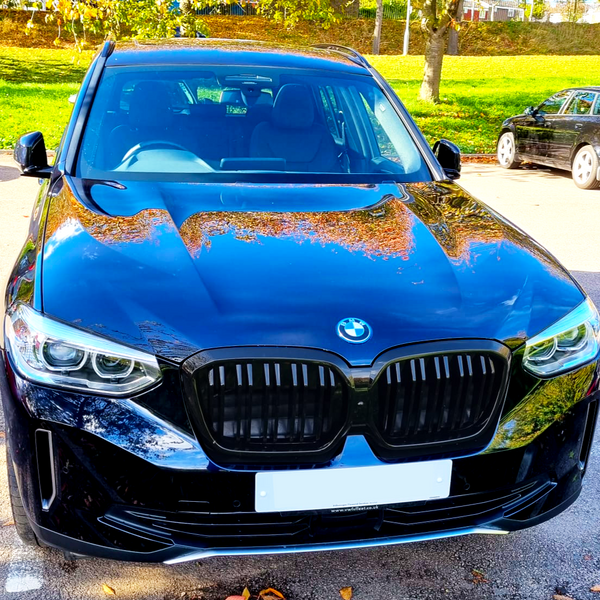 A black BMW SUV is parked on a sunlit street, with trees and grass visible in the background. The shiny car reflects nearby autumn foliage.