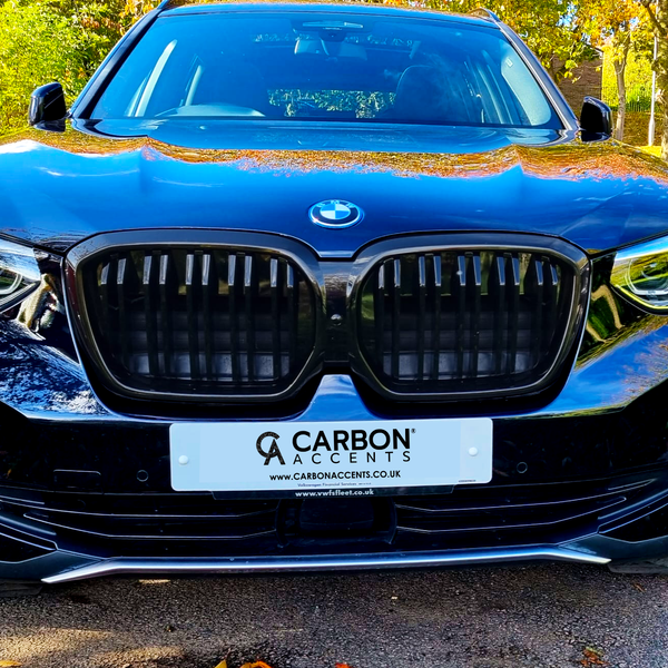A black BMW SUV is parked on a sunlit road, surrounded by trees. The front license plate reads: "CARBON ACCENTS" with websites "CARBONACCENTS.CO.UK" and "www.rvsfleet.co.uk."